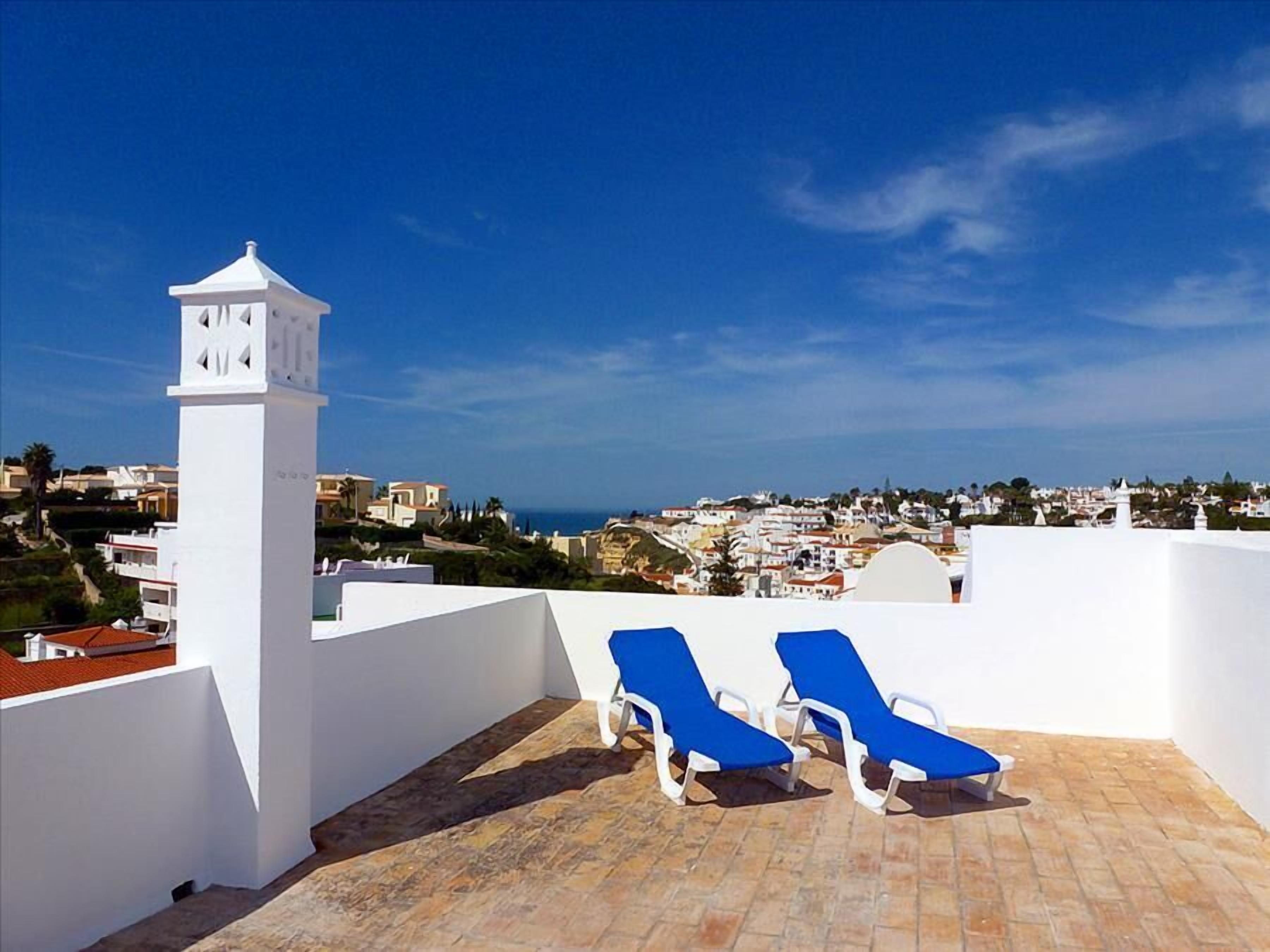 A rooftop terrace with two blue lounge chairs, a white tower, and a view of a coastal town.