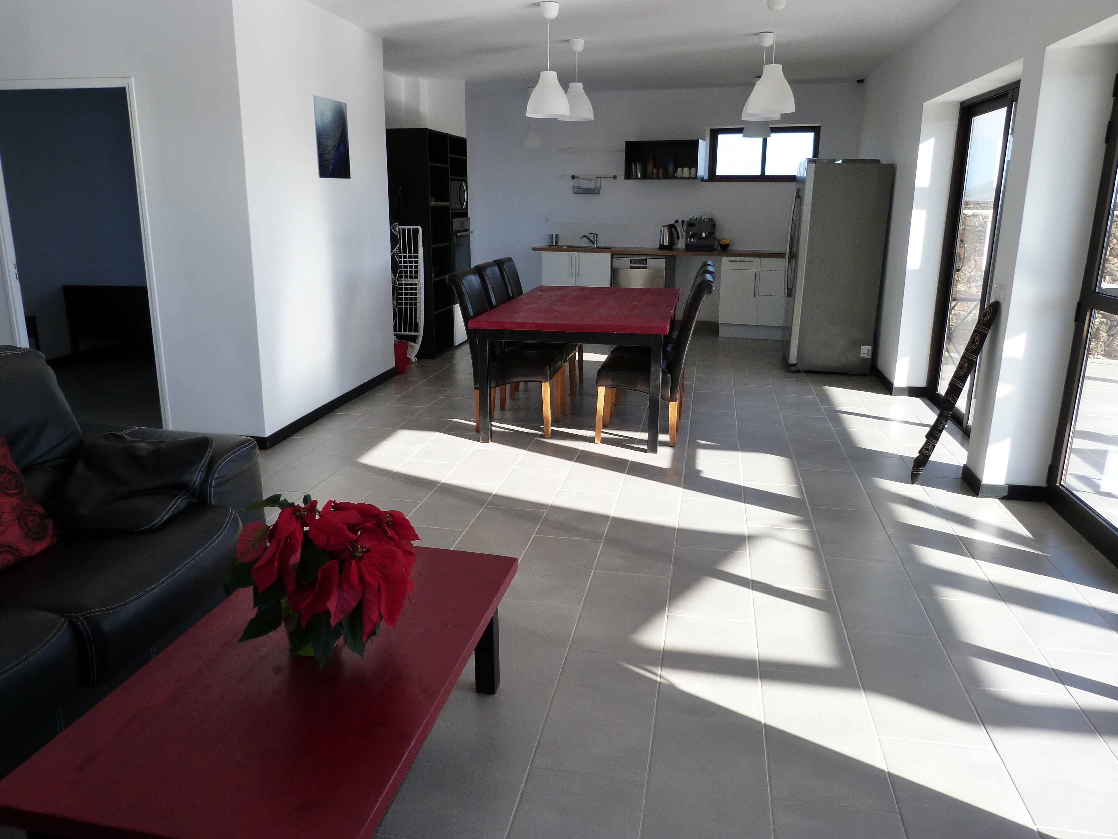 Una sala de estar moderna con una mesa roja y un sofá negro, un comedor con una mesa y sillas rojas, y una cocina con armarios blancos y luces colgantes.