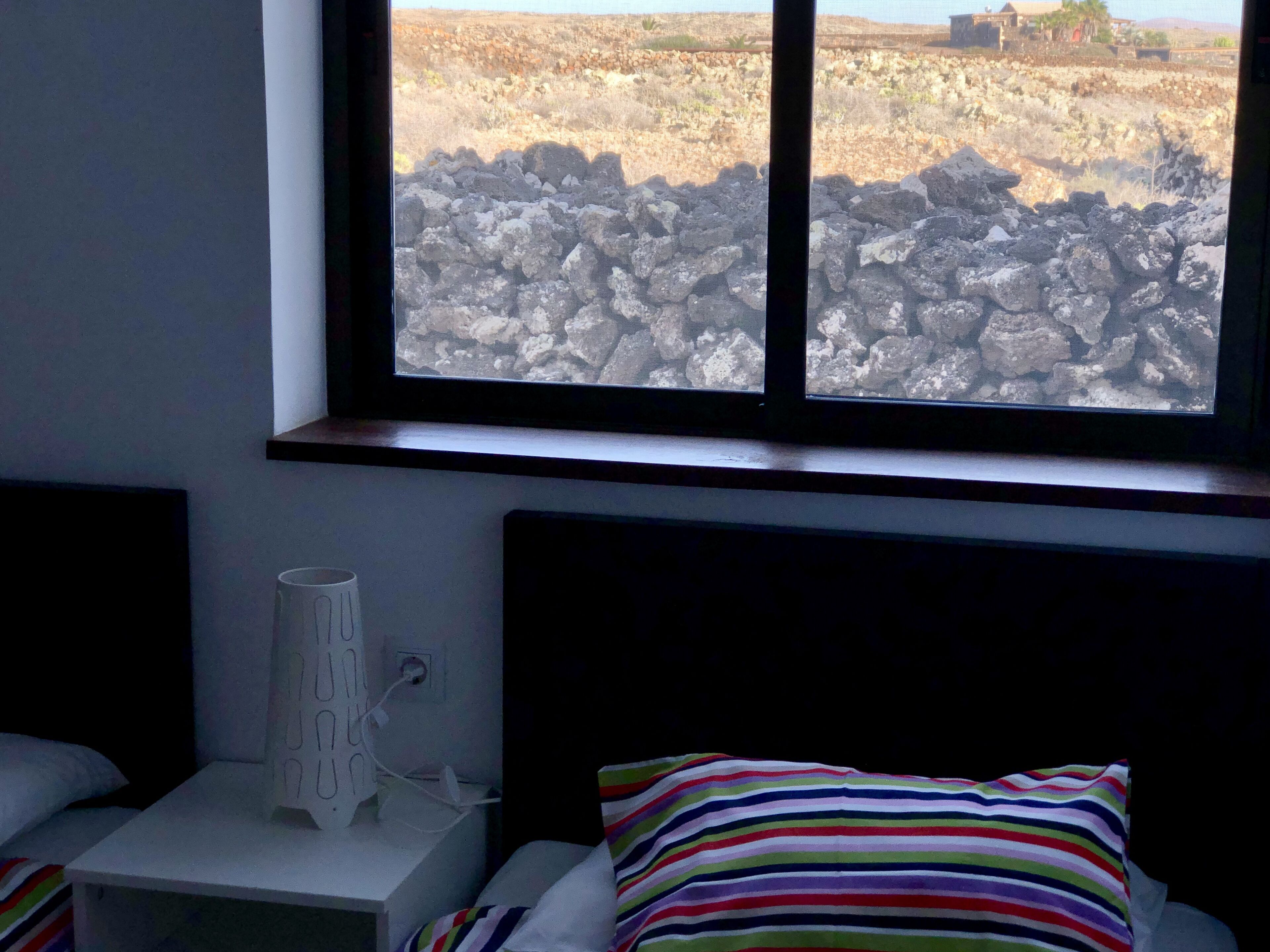 A bedroom with a bed, a bedside table with a lamp, and a window with a view of rocky terrain.