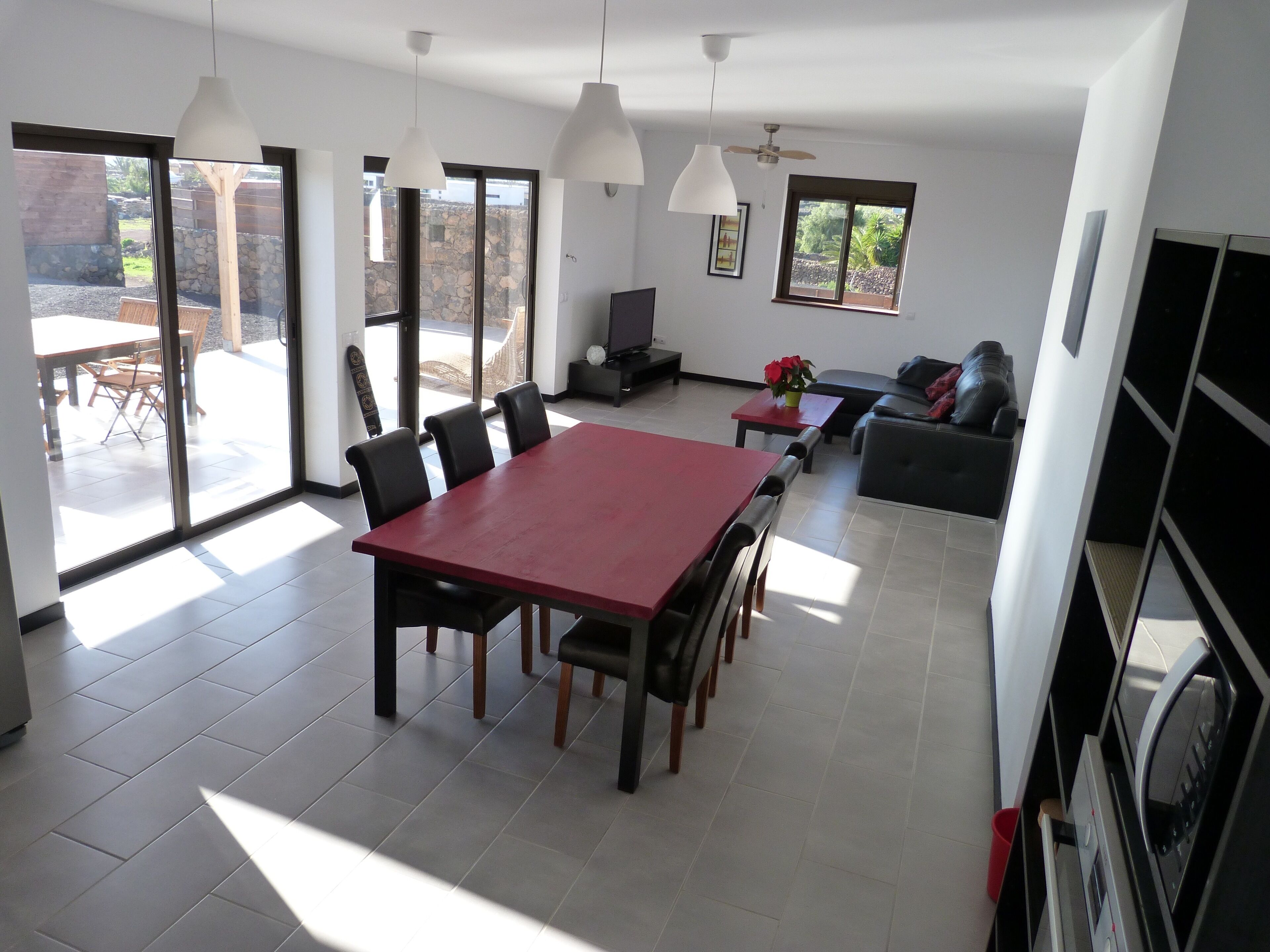 Een ruime woonkamer met een rode eettafel, zwarte stoelen en een grijze bank. Er zijn grote ramen die natuurlijk licht en uitzicht op de omgeving bieden.