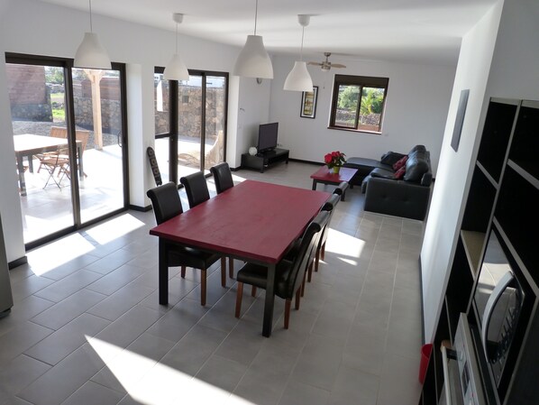 A spacious living area with a red dining table, black chairs, and a grey sofa. There are large windows providing natural light and a view of the outdoors.