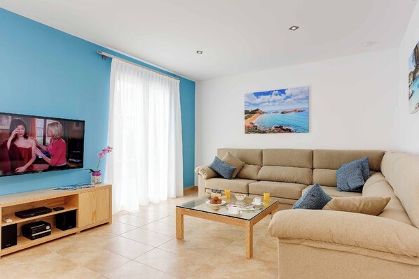 Un salón con sofá beige, una mesa de centro de vidrio, un televisor sobre un mueble de madera y un cuadro de una playa en la pared.