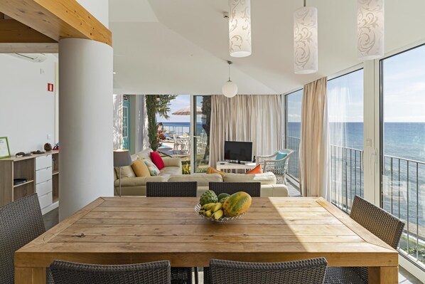 Un vaste espace de vie comprenant une table à manger en bois, des chaises en osier et une grande fenêtre donnant sur la mer.