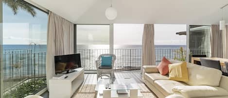 A modern living room with a large TV, comfortable seating, and a view of the sea.