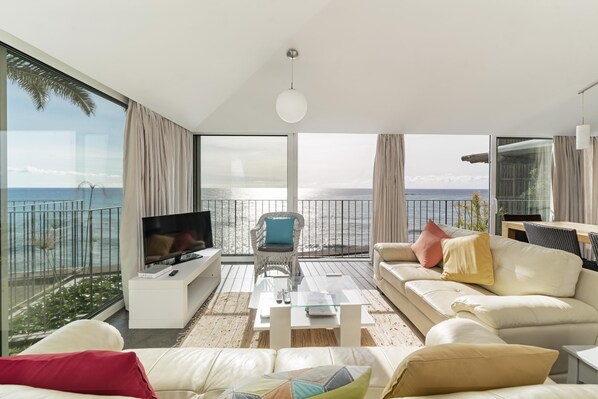 A modern living room with a large TV, comfortable seating, and a view of the sea.