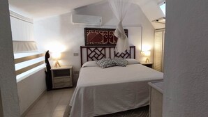 A bedroom with a bed, bedside lamps, a wall-mounted air conditioner, a decorative tapestry, and a window with curtains. - Villa with garden surrounded by greenery a stone's throw from the sea (Santa Margherita di Pula)