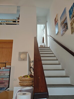 A staircase with a wooden handrail, a white wall with artwork, and a basket of fruit on the landing. - Villa with garden surrounded by greenery a stone's throw from the sea (Santa Margherita di Pula)