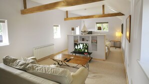 A spacious living room with a high ceiling, exposed wooden beams, a sofa, a coffee table, a television, and a fireplace. - The Stables at Partridge Lodge. (Bredfield, Woodbridge)
