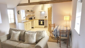 A cozy living room with a sofa, a dining table with chairs, a kitchen in the background, and a window providing natural light. - The Stables at Partridge Lodge. (Bredfield, Woodbridge)