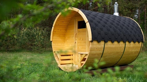 A wooden barrel-shaped cabin with a black roof, situated in a grassy area with trees in the background. - The Stables at Partridge Lodge. (Bredfield, Woodbridge)