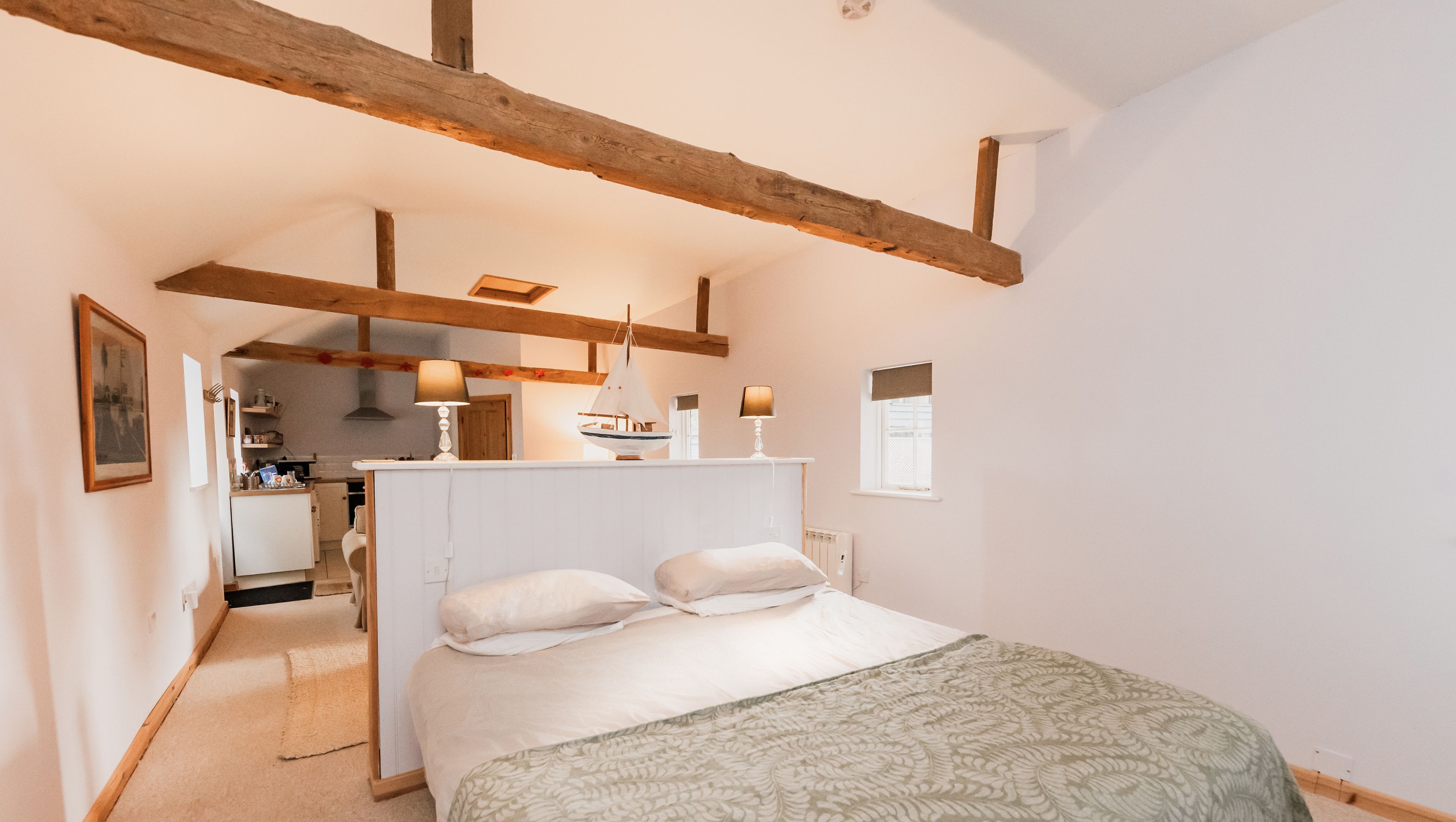Une chambre avec un lit, une poutre en bois au plafond, une petite table avec une lampe, et une vue sur la cuisine.