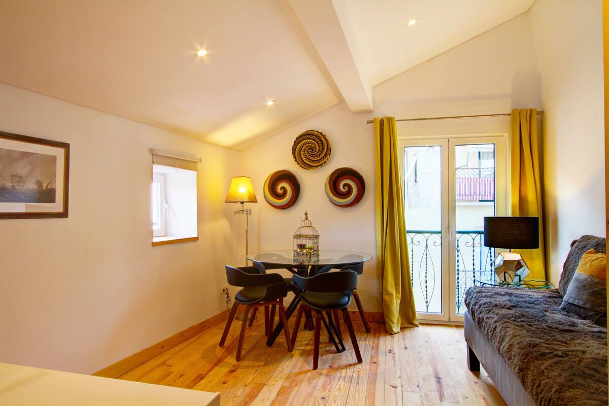 Una habitación con una mesa de comedor, sillas, una cama, un ventanal con cortinas y un balcón.