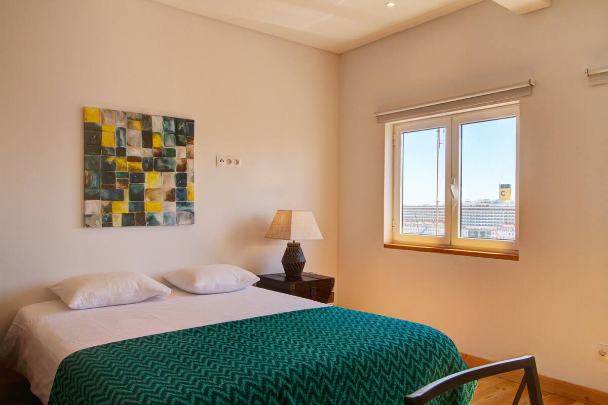 Una habitación de hotel con cama, mesita de noche, lámpara, ventana con vistas y un cuadro abstracto colorido en la pared.