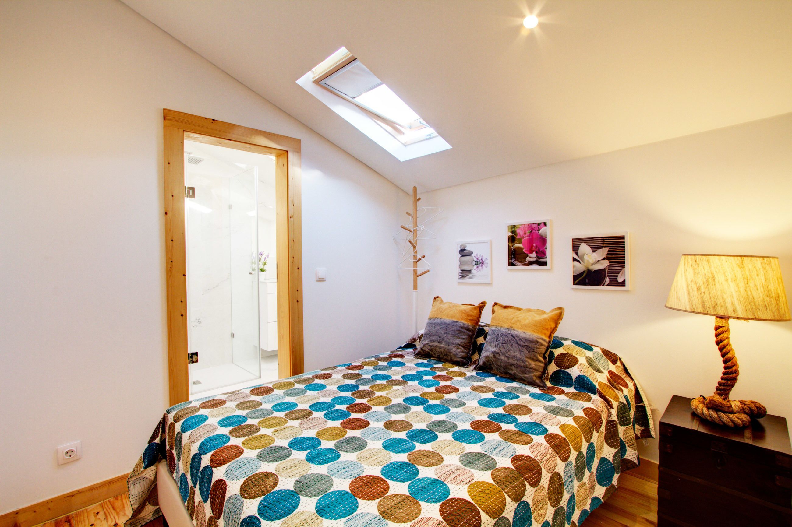 Une chambre avec un puits de lumière, un lit recouvert d’une couverture à pois, une lampe de chevet et une porte donnant sur une salle de bain.