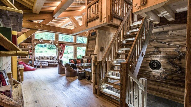 Een rustiek cabinteinterieur met een houten trap, een gezellige zithoek met kussens, en een boekenkast.