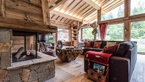 Een rustiek cabin interieur met een stenen openhaard, houten balken, een gezellige zithoek en een groot raam met uitzicht op de natuur.