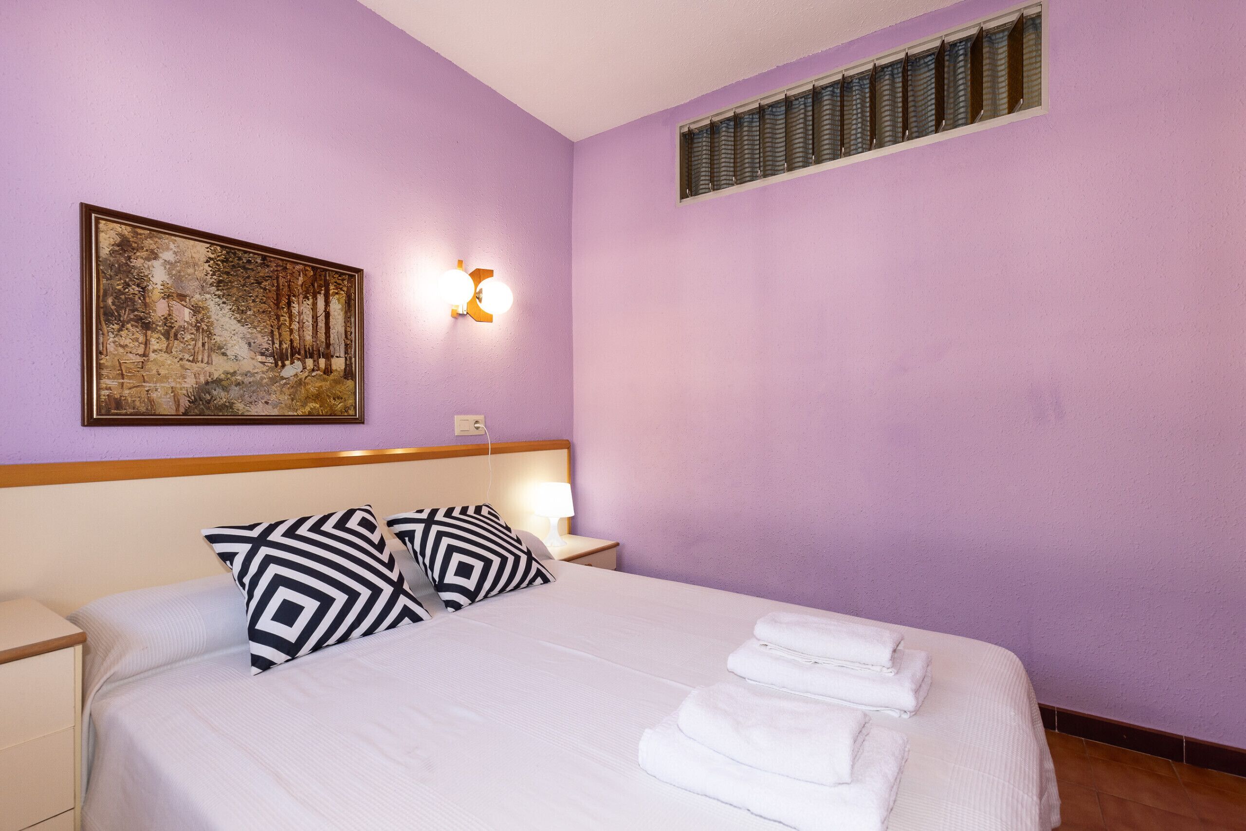 Habitación de hotel con cama, dos almohadas, mesita de noche, lámpara de pared y un cuadro.