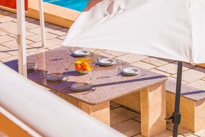 A table set outdoors with a white umbrella, plates, glasses, and a bowl of fruit. - Villa Zelda - PlusHolidays (Calpe)