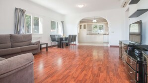 Amplia sala de estar con piso de madera, un sofá gris, una mesa de comedor con sillas y una cocina visible a través de un arco.