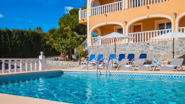 Piscina con sillones de descanso y sombrillas, rodeada por un muro de piedra y un edificio con balcones.