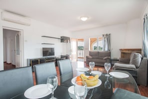 Un coin repas avec une table en verre, des chaises et une assiette de fruits. On aperçoit un canapé et un téléviseur au fond.