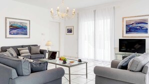 A modern living room with a grey sofa set, a glass coffee table, and a flat-screen TV. - Villa Polymnia (Kefalonia)