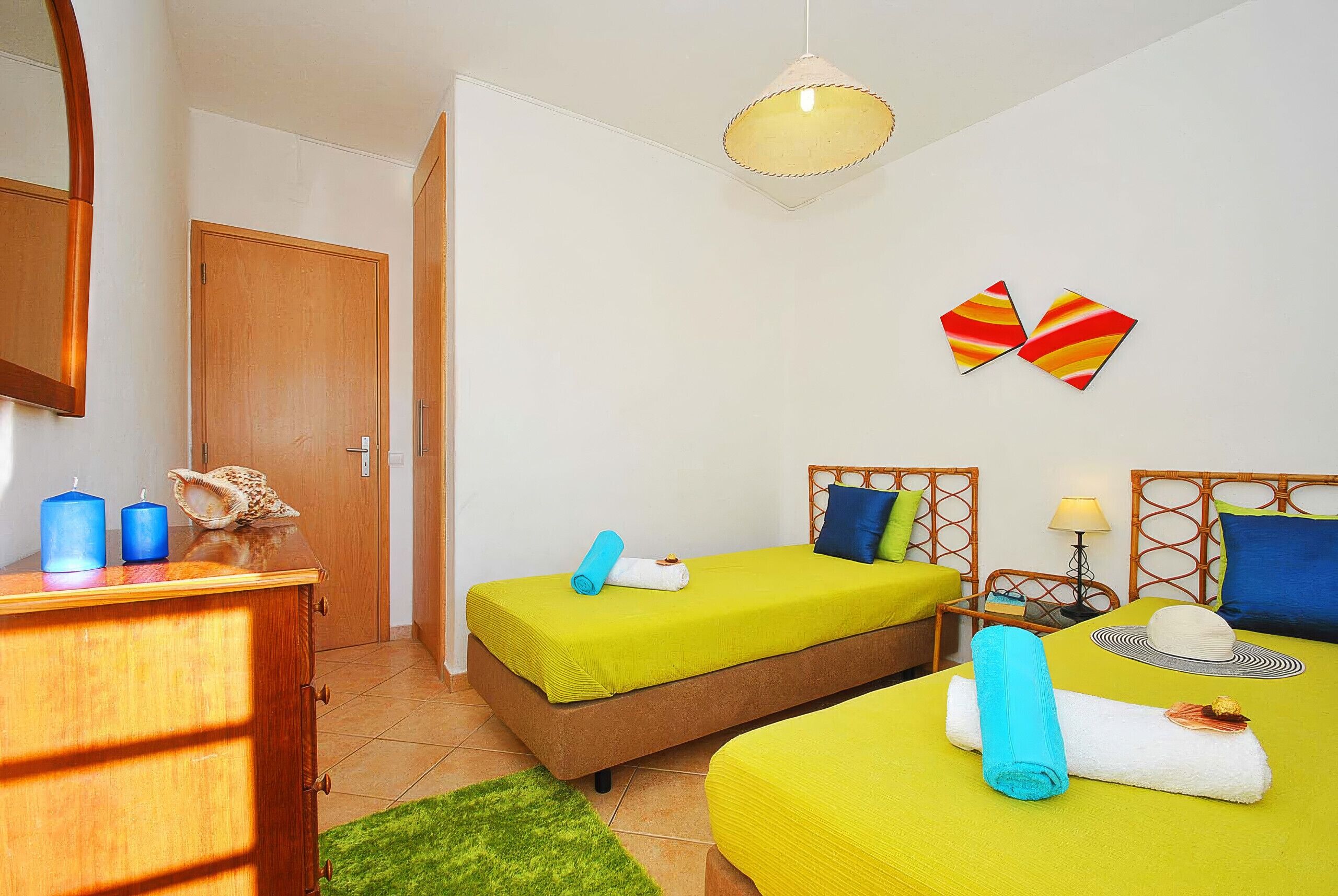 A room with two beds, a wooden dresser, a mirror, a green rug, and a wall with butterfly art.