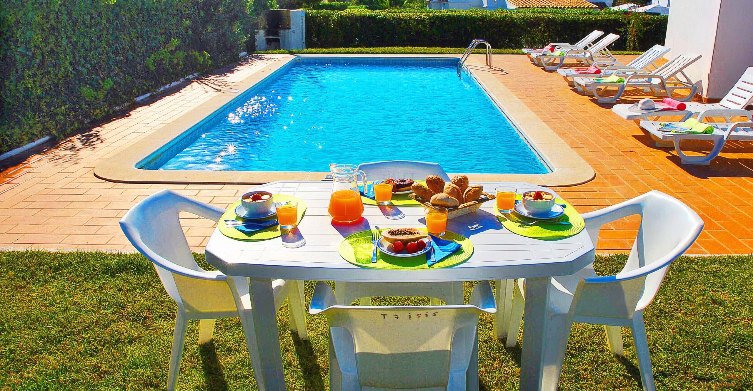 A poolside breakfast setup with a table, chairs, and a variety of food and drinks.
