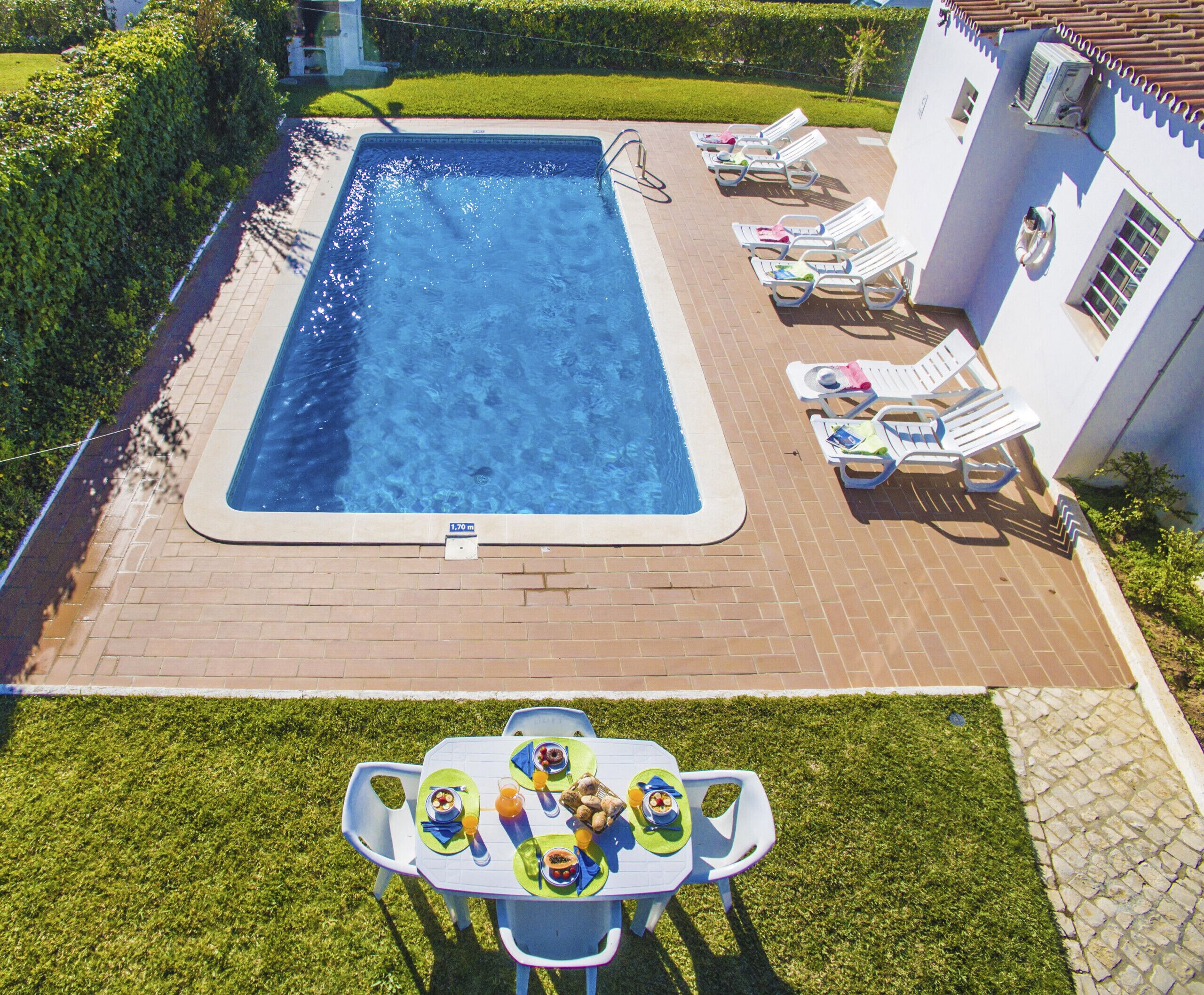 Une piscine rectangulaire d’une profondeur de 1,70 mètre, entourée de chaises longues et d’une table dressée pour un repas.