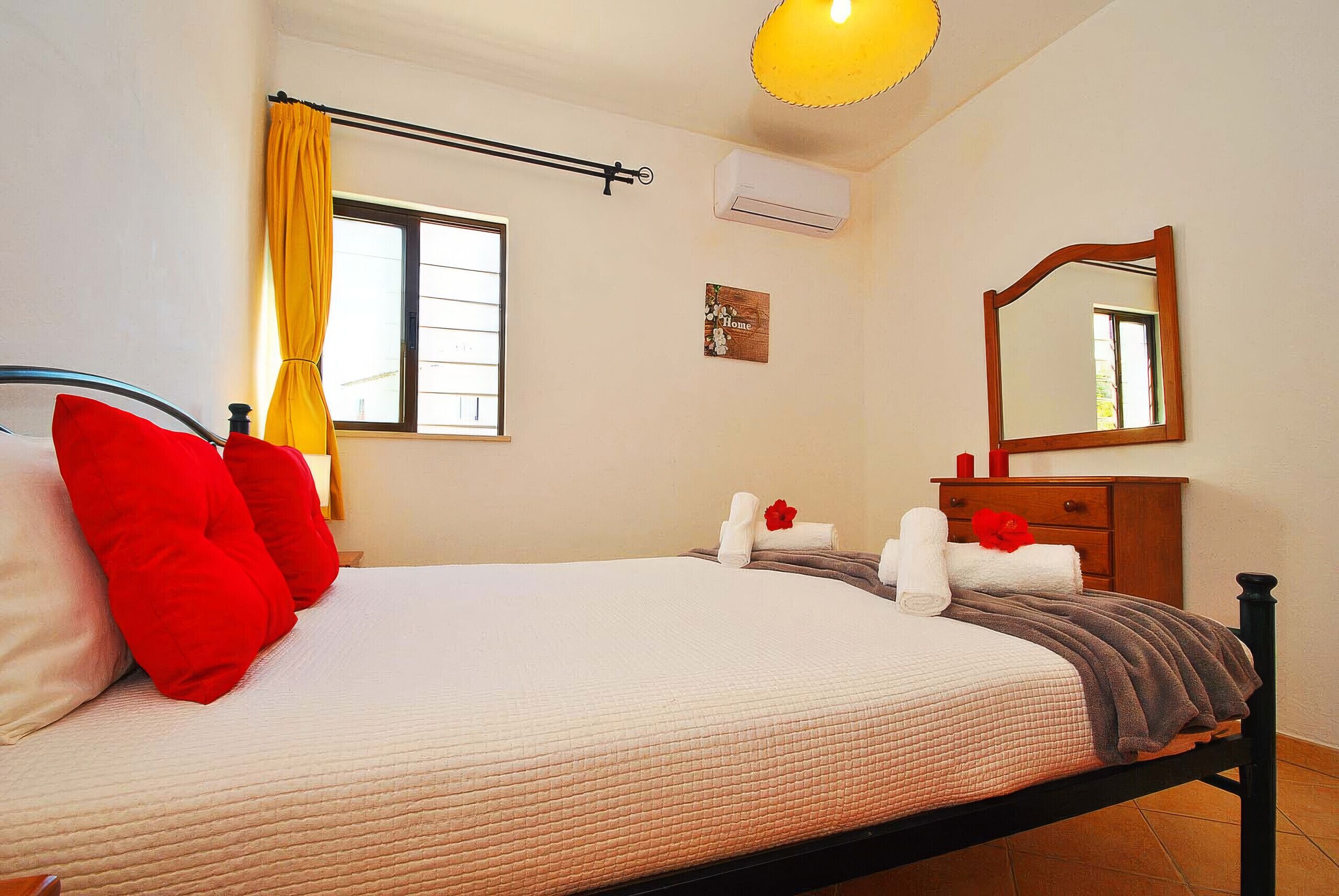A bedroom with a bed, a window with curtains, a mirror, and a bedside table with a lamp.