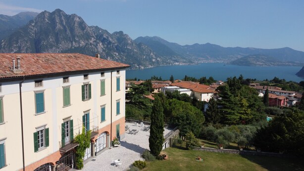 Un edificio con tejado de tejas y contraventanas verdes, un patio con mobiliario exterior y vistas a un lago y montañas al fondo.