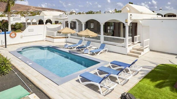 A swimming pool with lounge chairs and umbrellas, a white building with arches, and a clear sky.