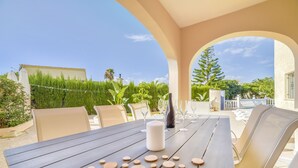 Ein Holztisch mit runden Untersetzern, eine weiße Kerze und Weingläser, draußen unter einer überdachten Terrasse mit beigen Wänden und blauem Himmel.