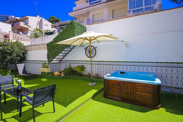 A backyard with artificial grass, a hot tub, a patio chair, and a table.