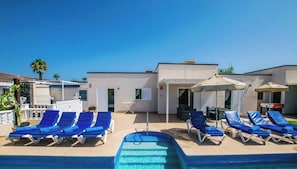 A pool area with lounge chairs and umbrellas, a clear sky, and a building with a flat roof.