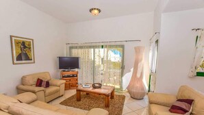 Ein Wohnzimmer mit beigefarbenen Sofas, einem Couchtisch, einem Fernseher und einem großen Fenster mit Vorhängen.