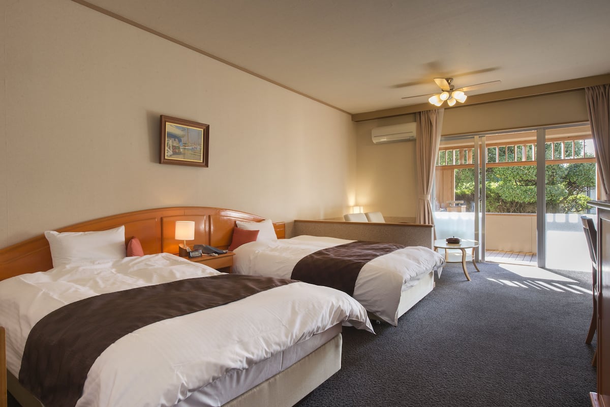 Habitación de hotel con dos camas, una mesita de noche, una lámpara, ventilador de techo, ventana con cortinas y una pequeña mesa.