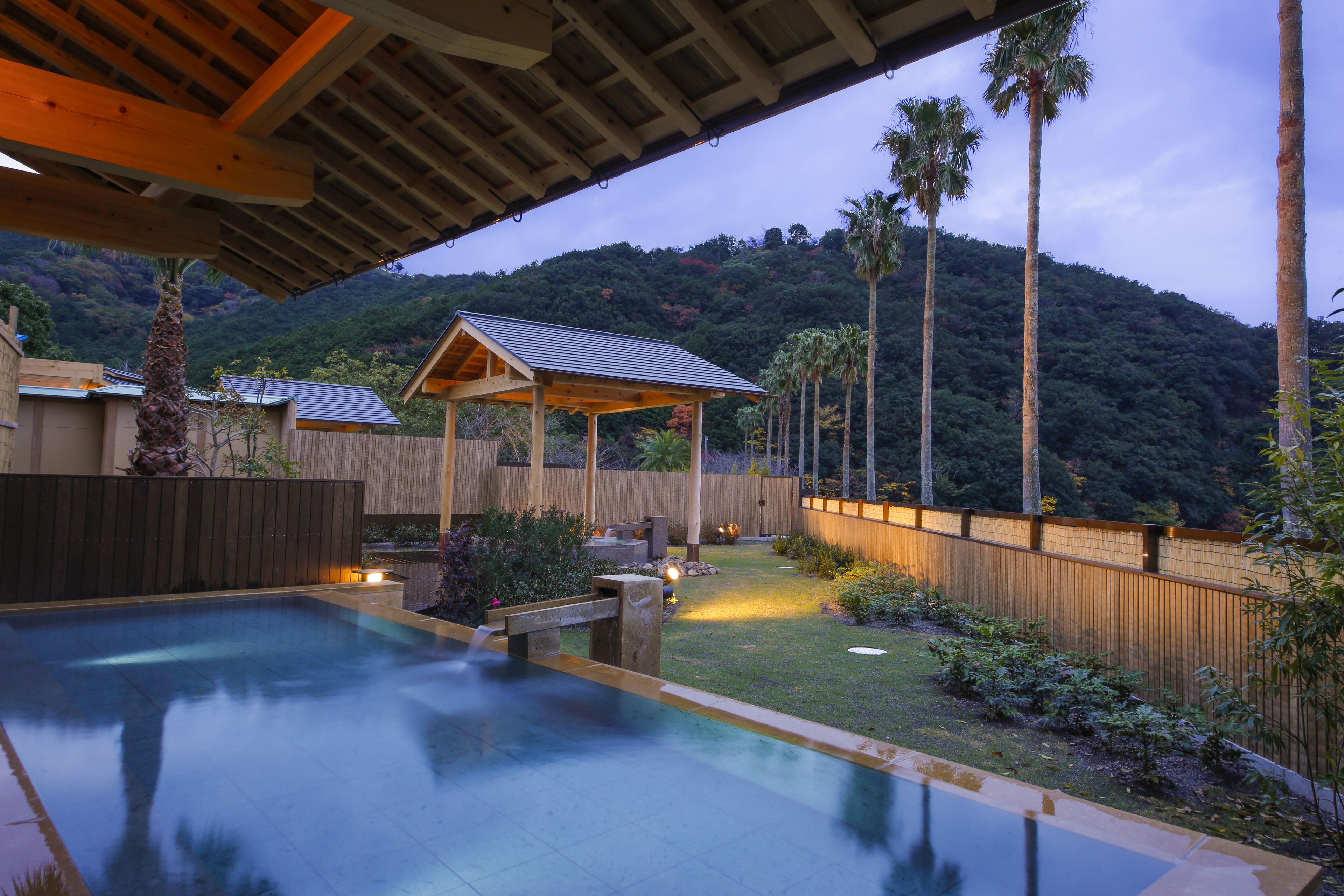 A pool area with a wooden deck, a covered seating area, and a view of a hillside.