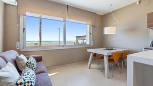 A modern living room with a large window overlooking a beach, a dining area with a white table and orange chairs, and a sofa with patterned cushions.