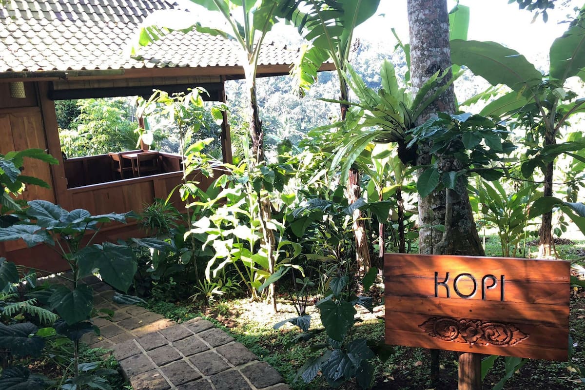 A wooden sign with the name 'KOPI' in a garden setting with various plants and a tiled roof structure in the background.