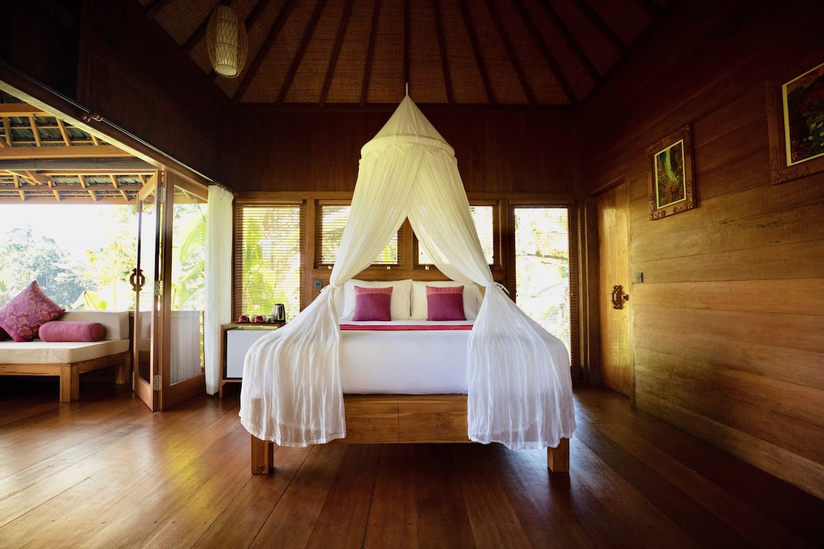 A wooden room with a bed under a white canopy, wooden floors, and a window with blinds.