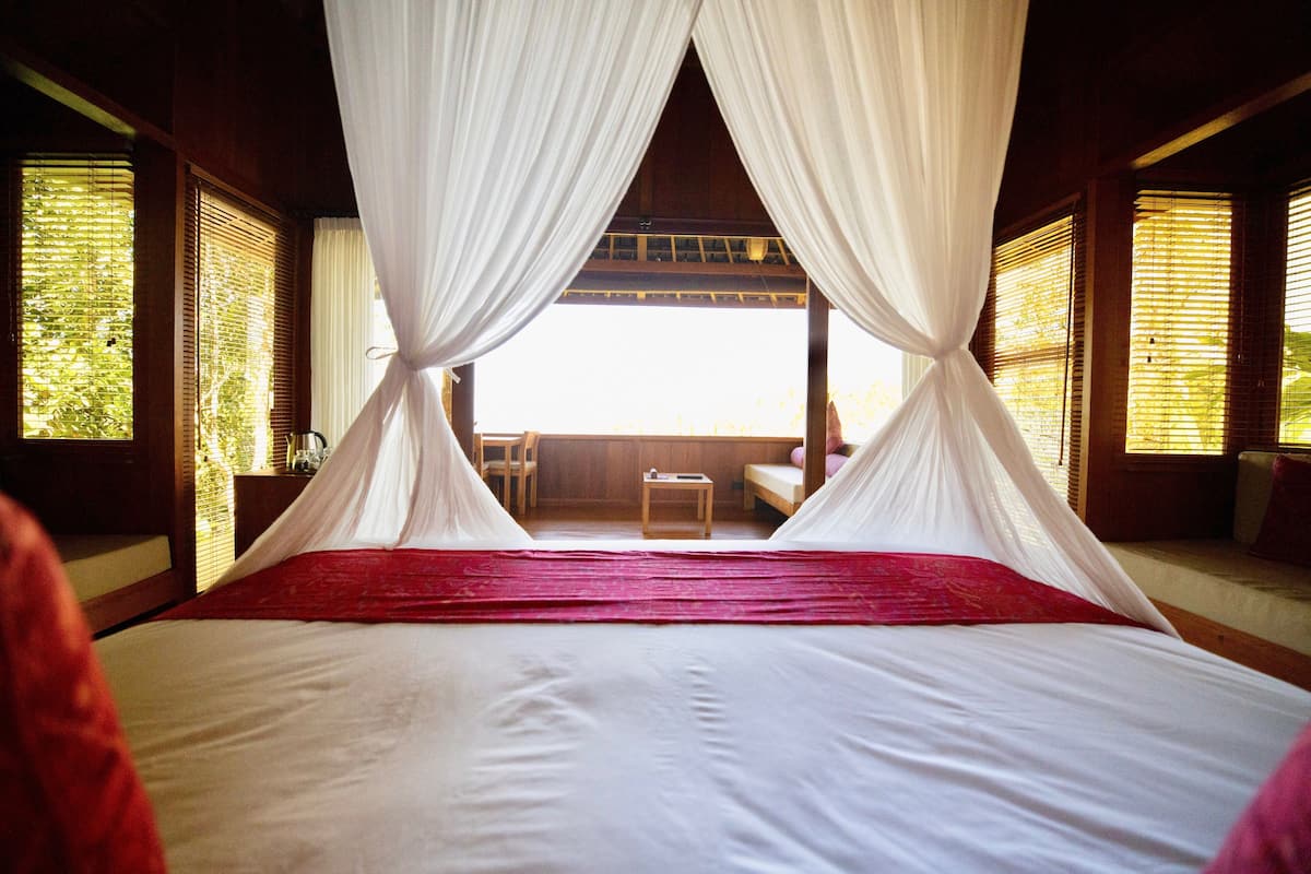 A bed with white linens and red accents, a wooden ceiling, and a view of the outdoors through windows with bamboo blinds.