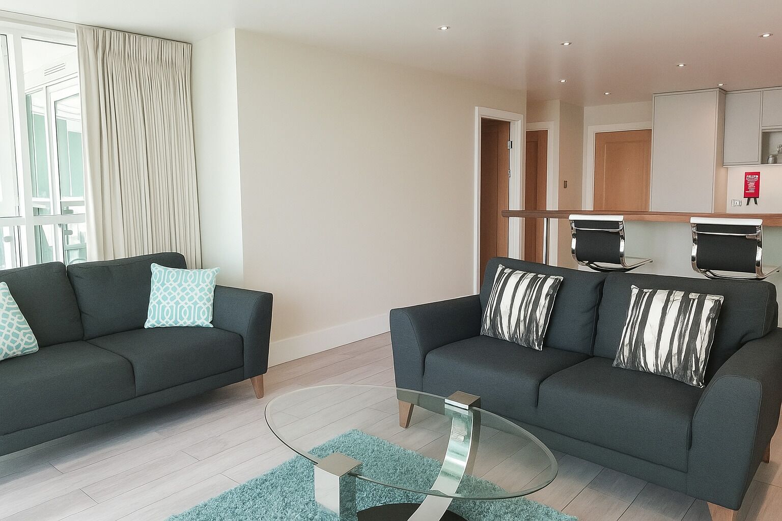 A modern living room with two sofas, a glass coffee table, and a kitchen area in the background.