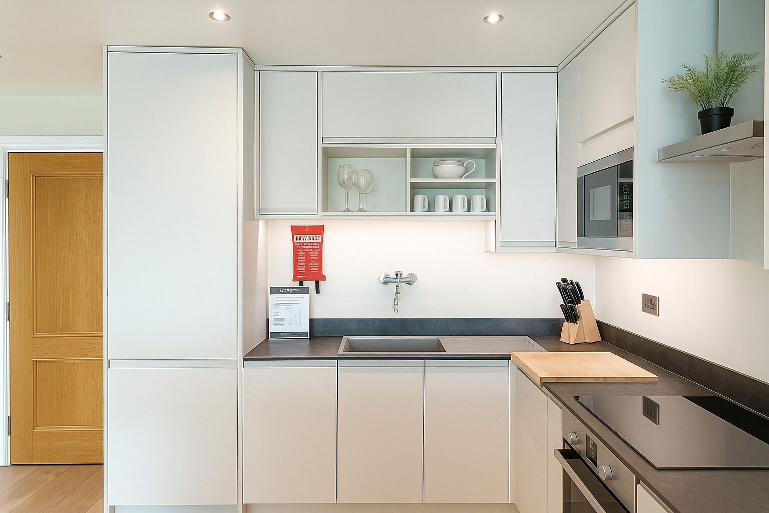 A modern kitchen with white cabinets, a stainless steel sink, and a microwave. There is a wooden door on the left and a plant on the right.