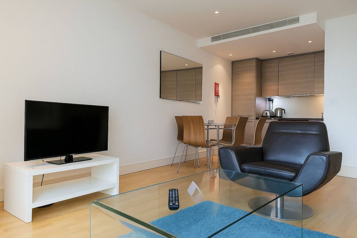 A modern living room with a flat-screen TV on a white console, a glass coffee table, a black leather sofa, a dining area with a table and chairs, and a kitchen area with cabinets and a kettle.