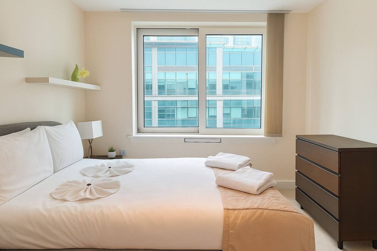 A hotel room with a large bed, a dresser, a window with a city view, and a small plant on the nightstand.