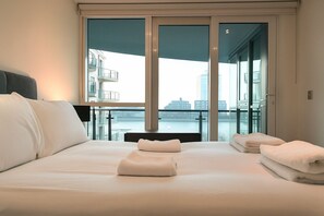 A hotel room with a large bed, two bedside tables, a view of a cityscape through floor-to-ceiling windows, and a balcony with a railing. - Still Life Vauxhall (London)