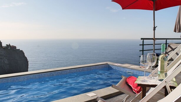 A poolside area with a view of the ocean, a red umbrella, and lounge chairs. - Perched above village, ocean views | Jardim Mar (Ponta do Sol)