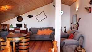 A cozy living room with a grey sofa, wooden barrels, and a small round table. - Perched above village, ocean views | Jardim Mar (Ponta do Sol)
