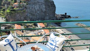A balcony with a view of a cliff and beach, featuring a table with a cushion and a decorative shell. - Perched above village, ocean views | Jardim Mar (Ponta do Sol)